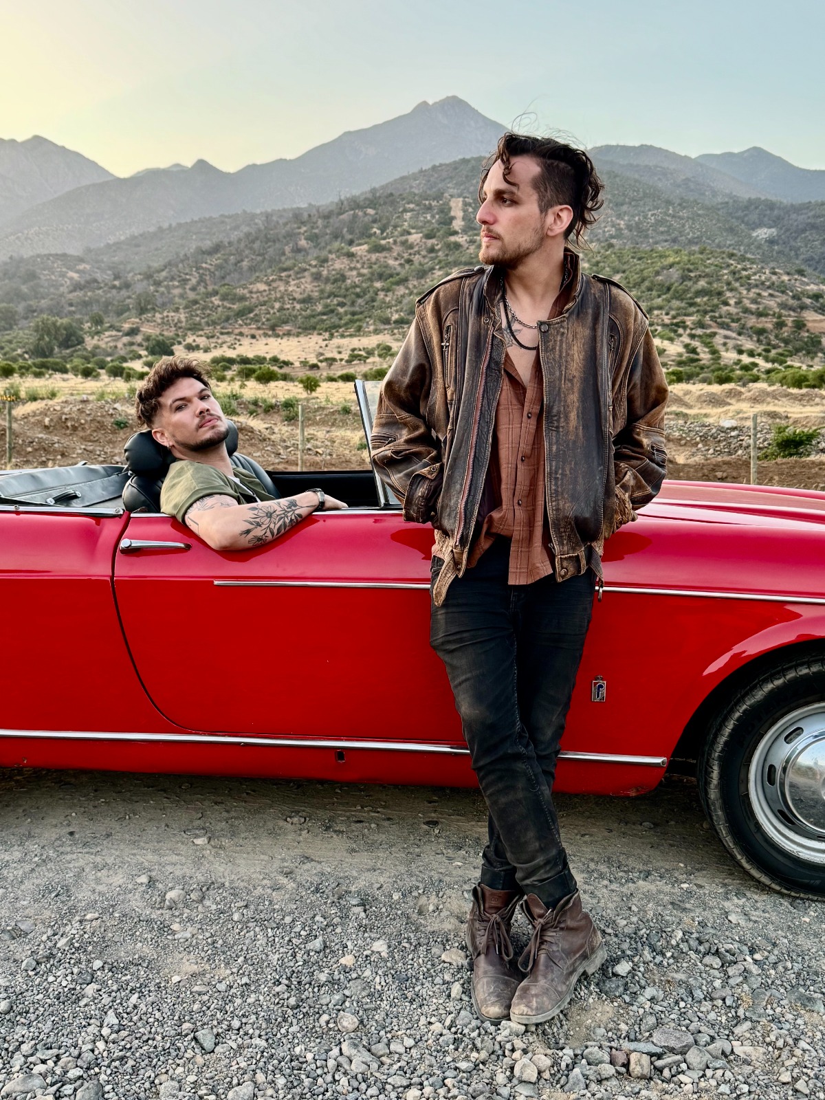 Diego y Chiris de Triciclo Parlante, estilo rock, junto a auto rojo convertible en paisaje montañoso. Álbum Re:nacer.