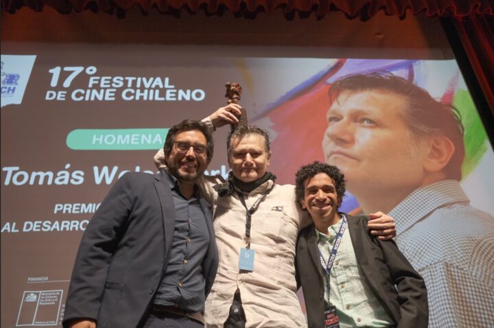 Tomás Welss, homenajeado en 17° FECICH por Aporte a la Animación Nacional, recibe premio junto a dos hombres. Pantalla con su retrato.