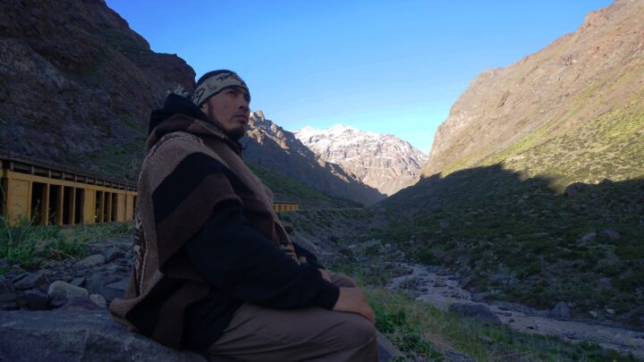 Neculman, rapero mapuche, con poncho andino en la Cordillera de Los Andes. Medita ante montañas nevadas. Videoclip 'Andes'.