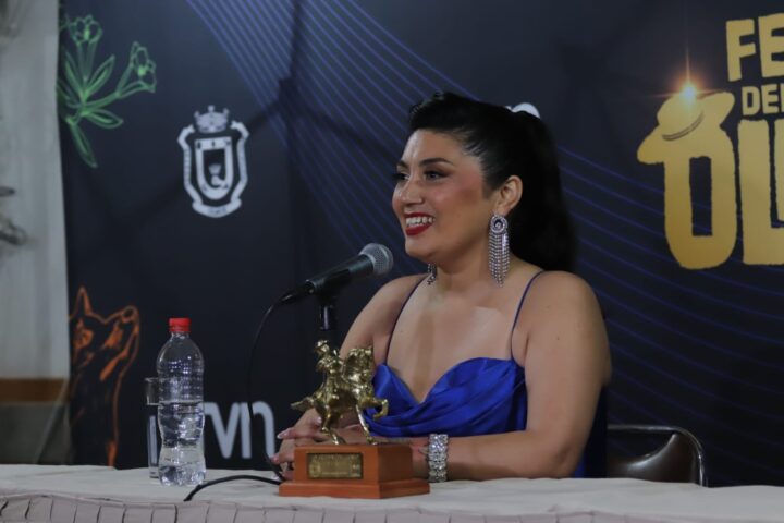 Mujer con vestido azul y aros largos sonríe en conferencia de prensa del Festival del Huaso de Olmué junto al Guitarpín Dorado.