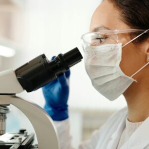 Investigadora en laboratorio, con mascarilla y gafas, usa microscopio. Representa el futuro de la mujer en STEM.