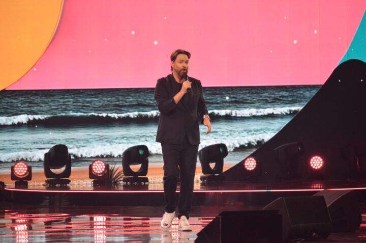 George Harris, comediante venezolano, en el escenario con micrófono. Fondo de playa y cielo rosa. Su show fue polémico en Viña.