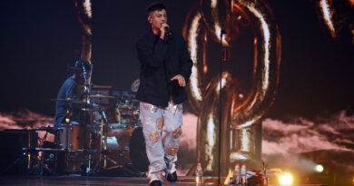 Duki, ícono del trap, en Viña 2025. Canta con micrófono, tatuajes faciales, pantalón estampado, escenario con cadenas.