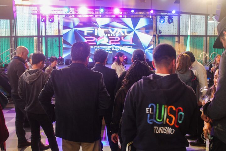 Público en FITVAL 2024, Feria Internacional de Turismo Valparaíso Región. Pantalla con logo y luces. Persona con polerón 'El Quisco Turismo'.