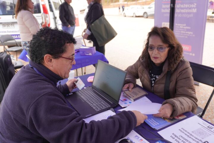 Funcionario IPS ChileAtiende asiste a adulta mayor con solicitud PGU en mesa con laptop y documentos.