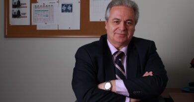 Profesional con traje azul, camisa rosa y corbata, sentado en escritorio con brazos cruzados. Fondo: tablón de anuncios con horarios y logos Radio UCentral.