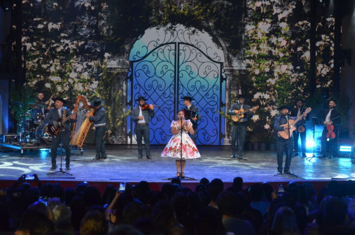 Agrupación folclórica "Silvanita y los del Quincho" en escenario del Festival del Huaso de Olmué. Músicos con arpa, guitarras, violín. Fondo digital floral y portal.