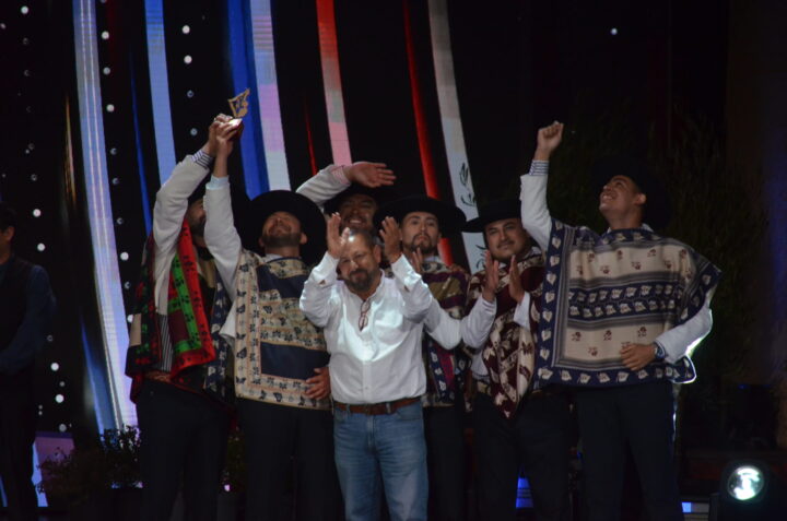 Grupo de artistas folclóricos celebrando en escenario, uno levanta un trofeo. Visten ponchos y sombreros. Premiados por su música.