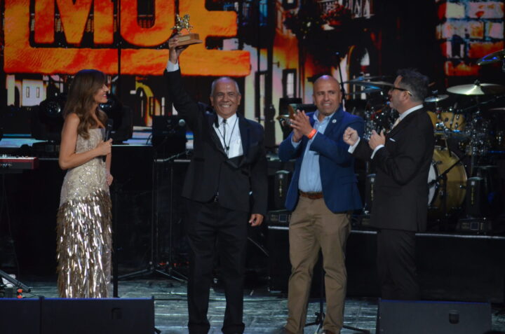 Hombre levanta trofeo de caballo dorado en escenario. Presentadora y dos hombres aplauden en gala musical. Fondo con pantalla.
