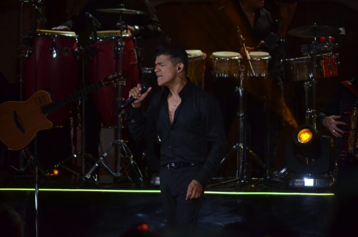 Rey de la Cumbia Chilena canta apasionado en concierto de Olmué. De negro, micrófono en mano, congas y guitarra al fondo.