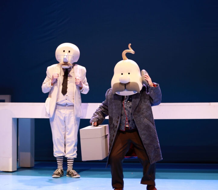 Actores enmascarados en escena teatral: uno de blanco, otro con maleta. Representación de la programación de verano.