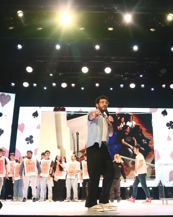 Edo Caroe en vivo en escenario, apunta. Artistas y pantalla con iconos de cartas en recinto patrimonial.