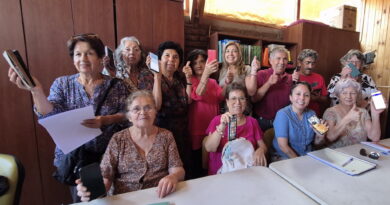 Grupo de adultos y jóvenes sonríen con smartphones y pulgares arriba en taller contra brecha tecnológica para salud y redes familiares.