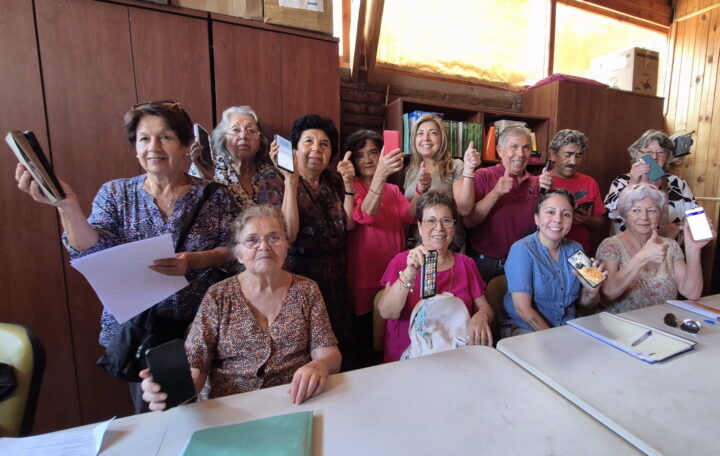 Grupo de adultos y jóvenes sonríen con smartphones y pulgares arriba en taller contra brecha tecnológica para salud y redes familiares.