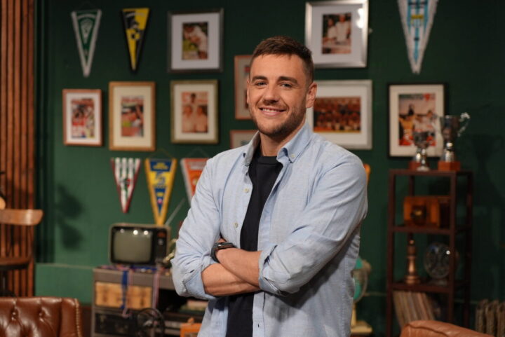 Sonriente Gonzalo Fouillioux en set deportivo. Viste camisa celeste, brazos cruzados. Fondo verde con fotos, trofeos y banderines.