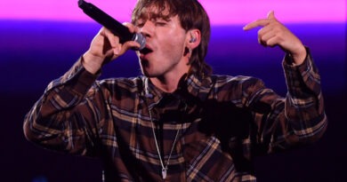 Paulo Londra canta enérgicamente en concierto. Viste camisa a cuadros, sostiene micrófono, señalando al público, con luces moradas de fondo.