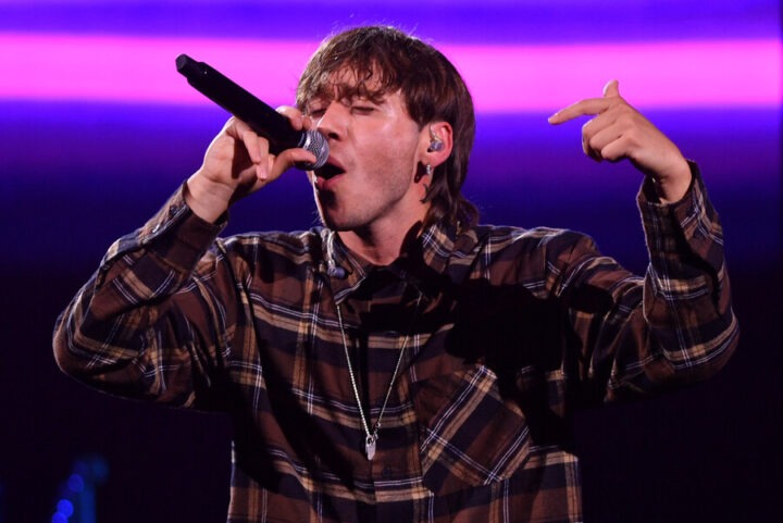 Paulo Londra canta enérgicamente en concierto. Viste camisa a cuadros, sostiene micrófono, señalando al público, con luces moradas de fondo.