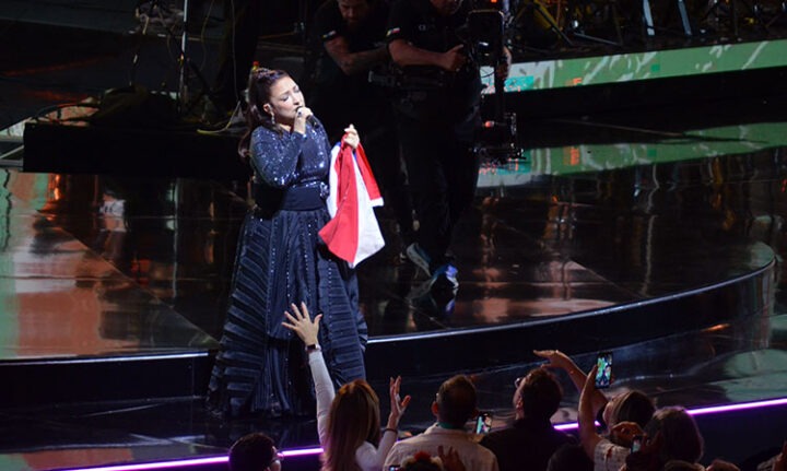 Gloria Estefan en el escenario de Viña del Mar, cantando con vestido negro, sosteniendo bandera chilena, público emocionado.