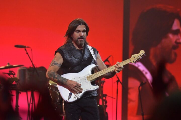 Juanes con chaleco, tatuajes, tocando guitarra eléctrica blanca en escenario rojo con proyección.