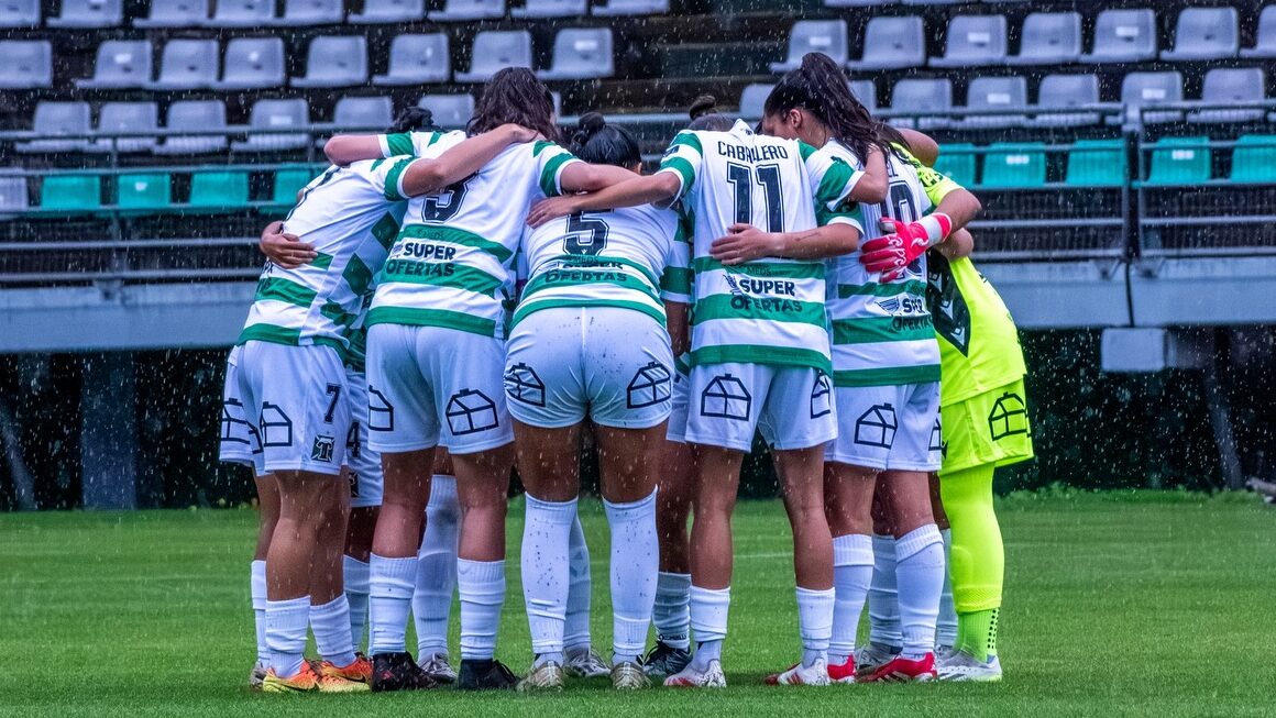 Plantel de fútbol femenino reunido en el campo de juego bajo la lluvia.