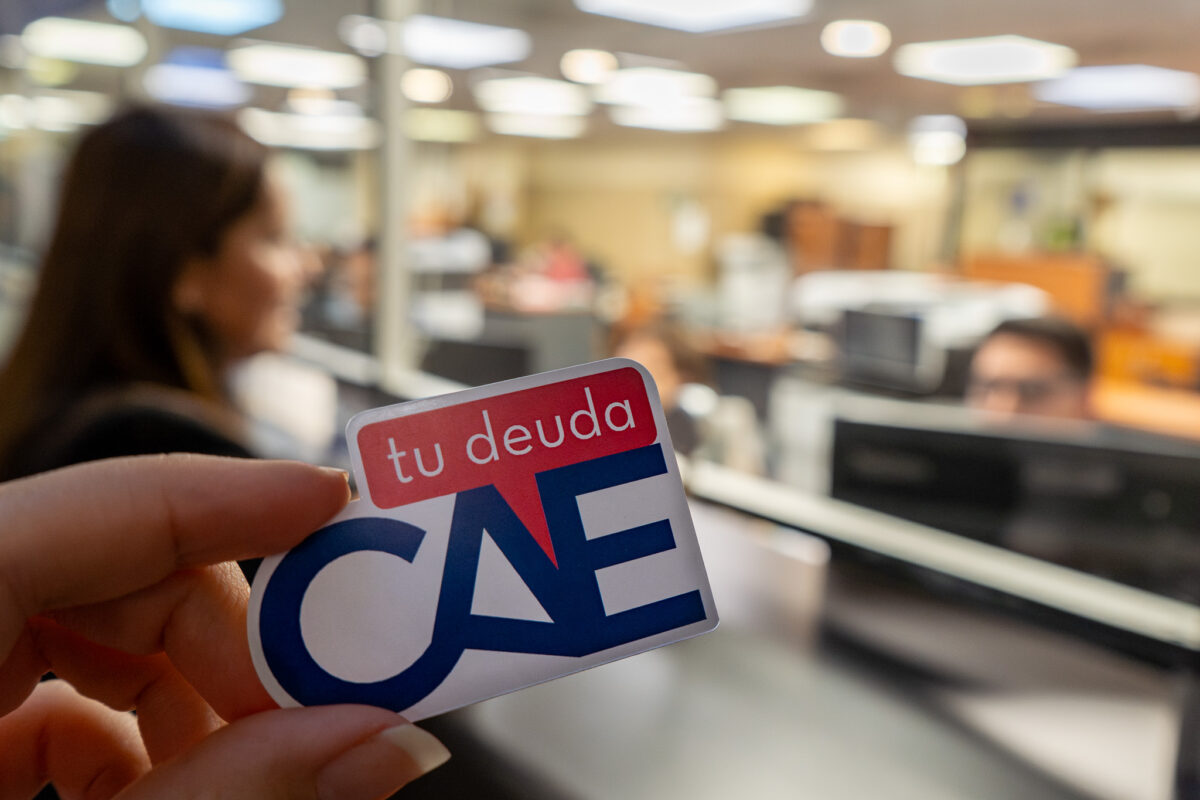 Una persona sosteniendo un sticker con el texto 'tu deuda CAE' en el contexto de una oficina.