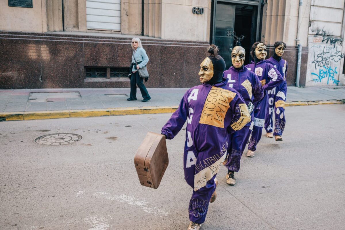 Grupo de personas con máscaras doradas y trajes morados caminando en fila por una calle.