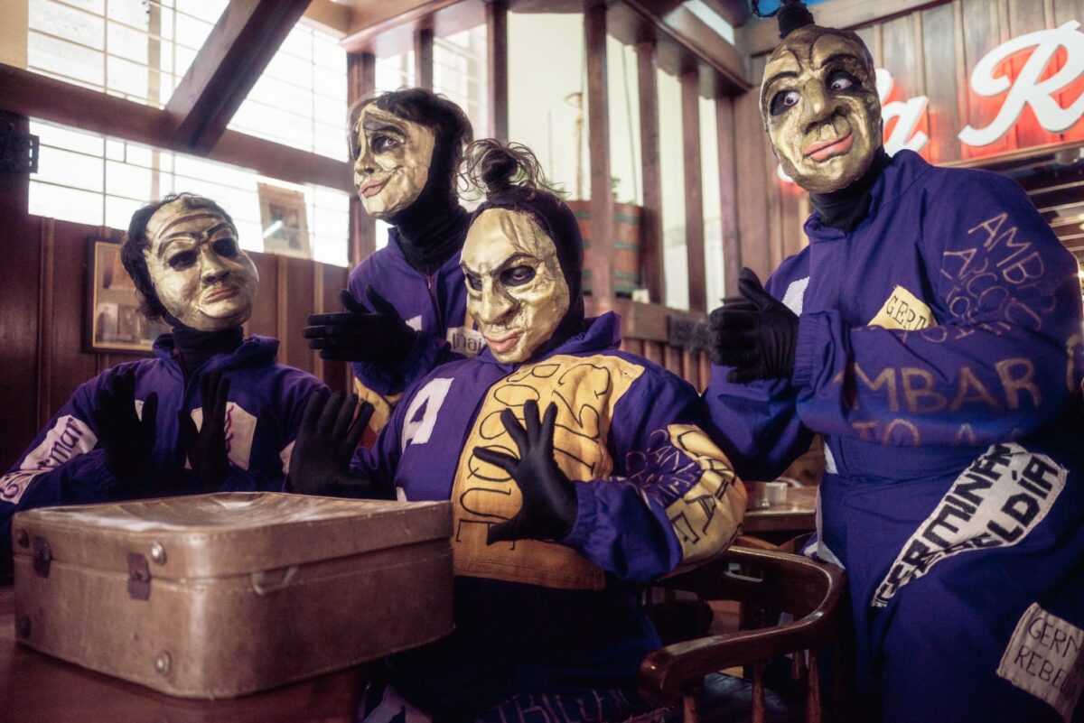 Cuatro personas con máscaras doradas y trajes morados sentadas alrededor de una mesa, una de ellas sosteniendo un balón.
