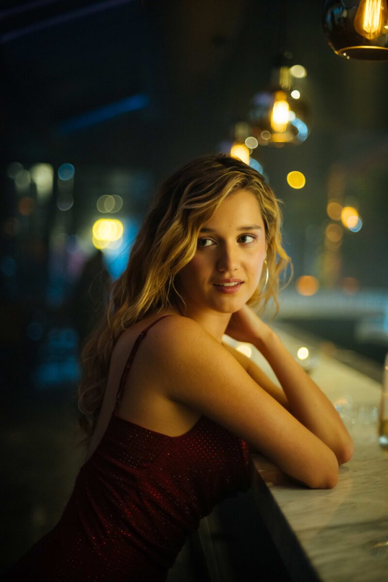 Una joven rubia con un vestido brillante apoyada en una barra con luces de fondo.