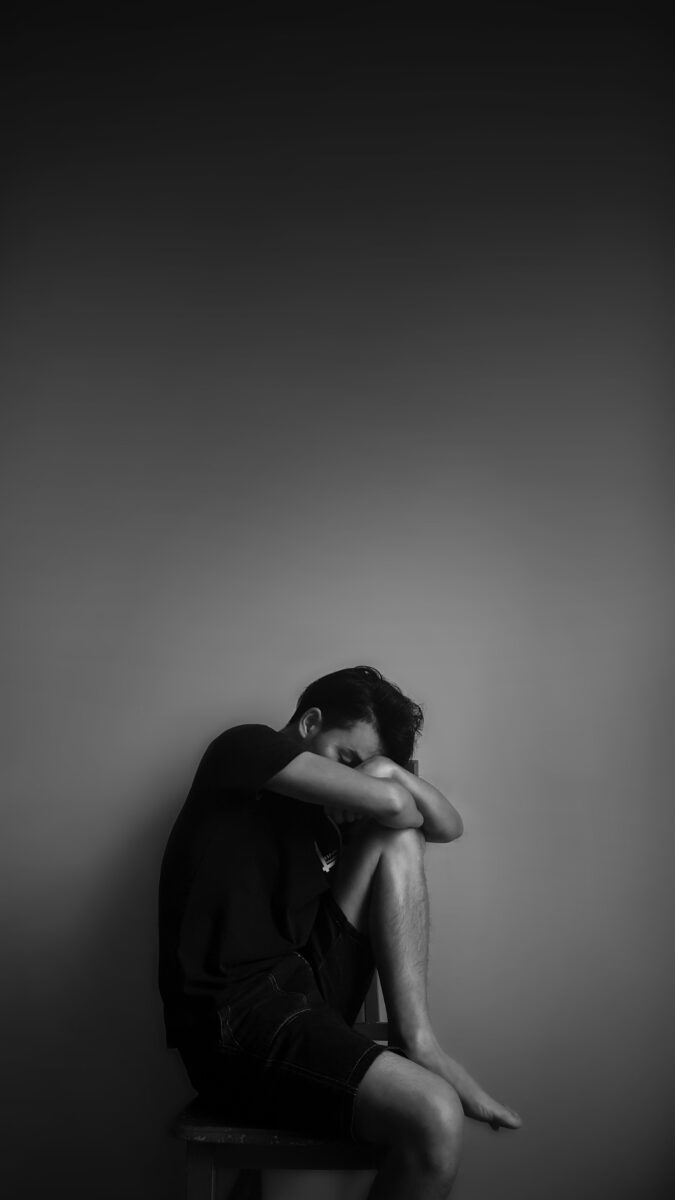 Hombre joven sentado encogido con la cabeza entre las rodillas, en blanco y negro.
