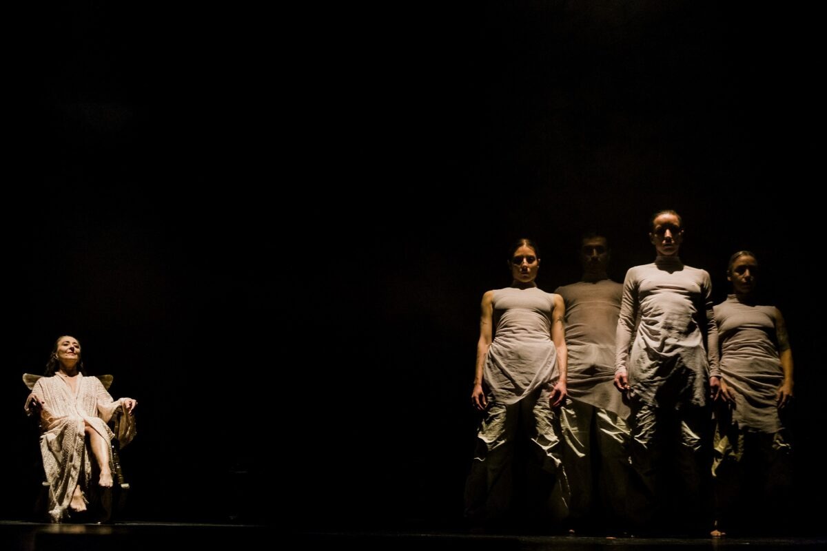 Escena de la obra de danza THITA con una intérprete sentada y un grupo de bailarines al fondo en la penumbra.