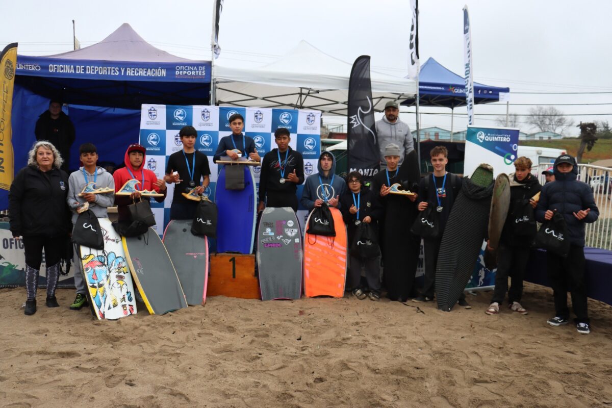 Grupo de jóvenes deportistas posando con sus trofeos y tablas en la arena de Playa El Bato, Quintero.