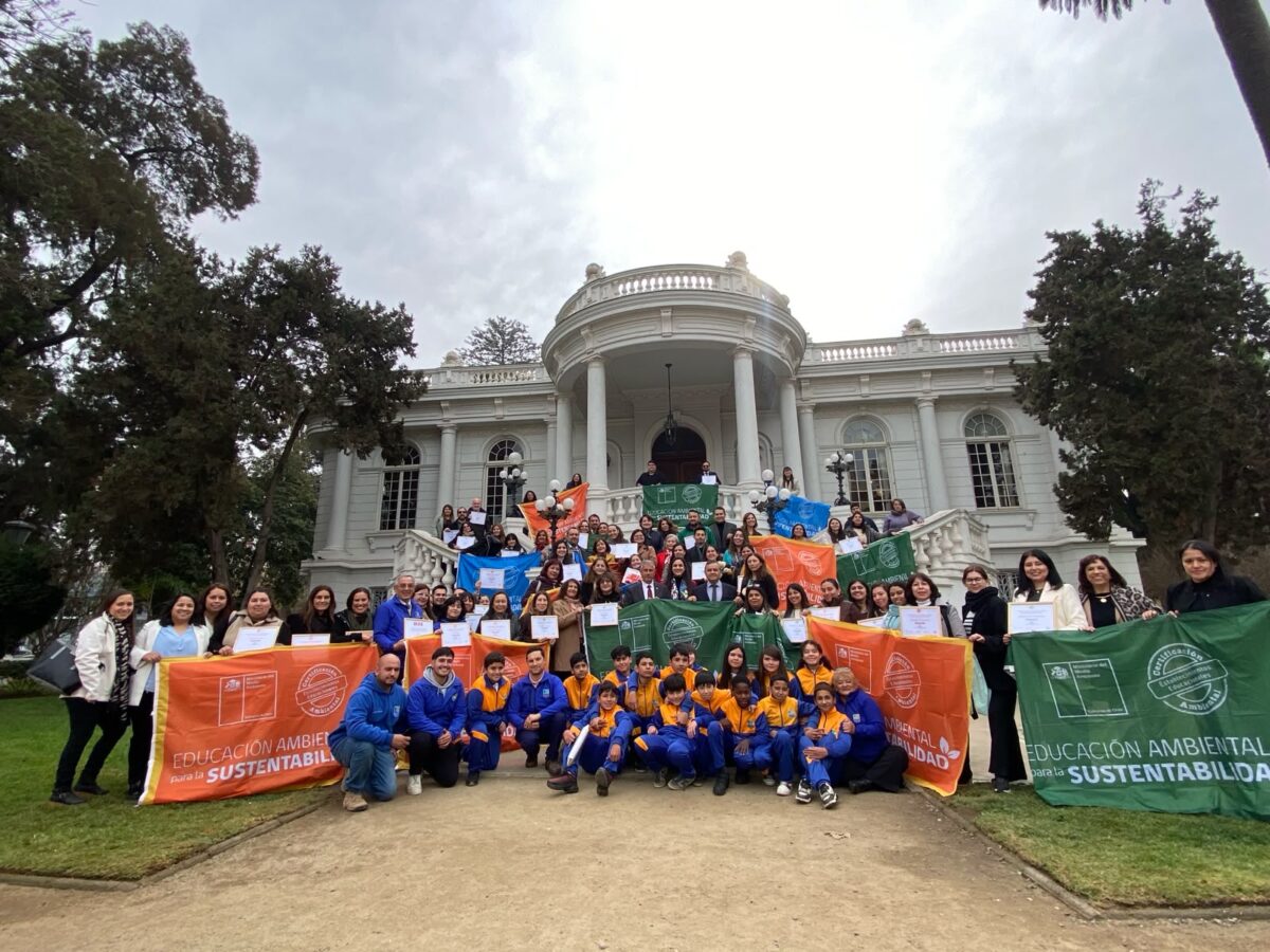Grupo de personas, entre estudiantes y adultos, posando con banderas y certificados de educación ambiental frente a un edificio.