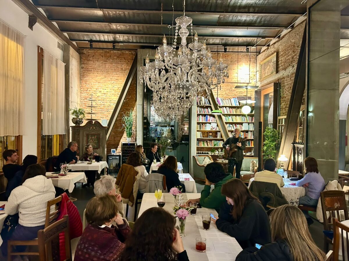 Interior del restaurant Da Mafalda, mostrando a comensales y un músico tocando la guitarra en un ambiente acogedor con un gran candelabro y estanterías de libros.