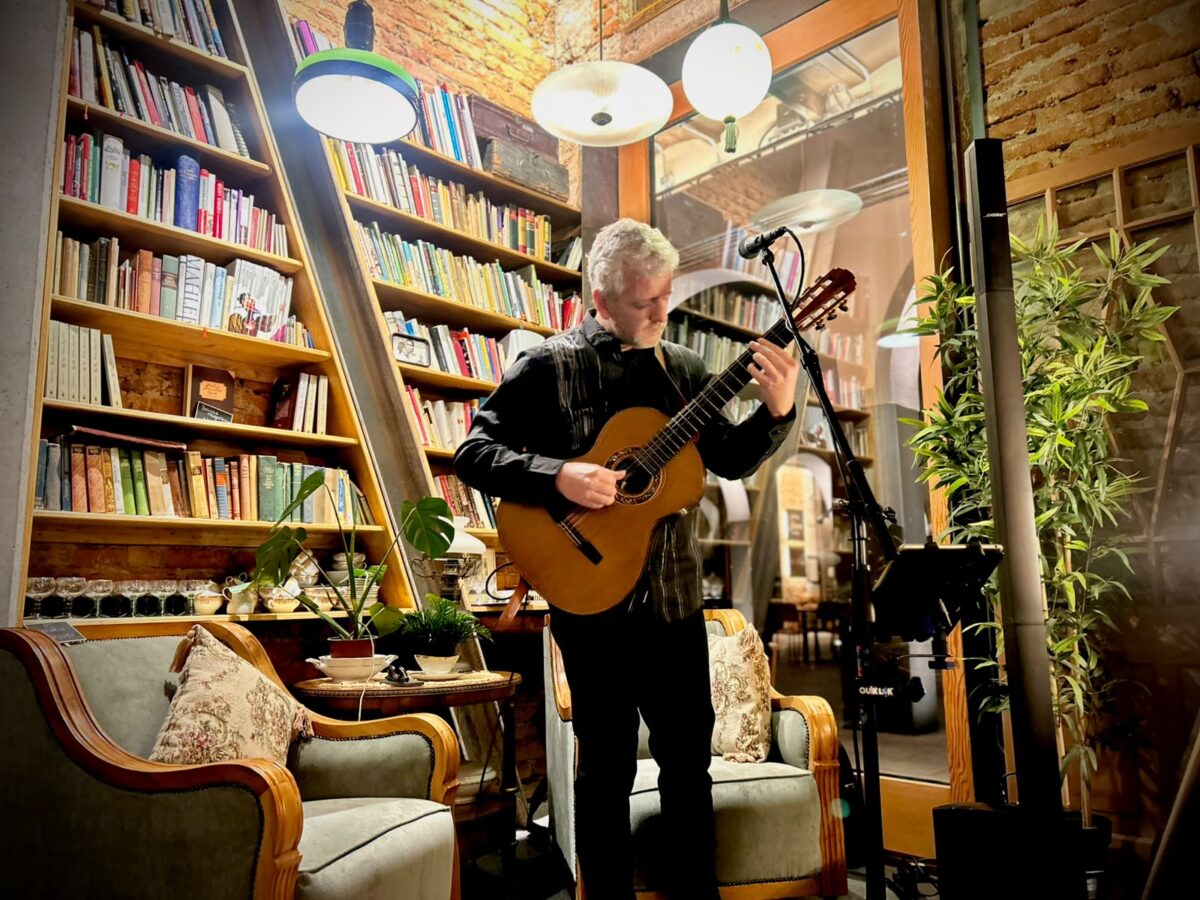 Músico tocando la guitarra en el escenario de Da Mafalda, con una estantería de libros de fondo.