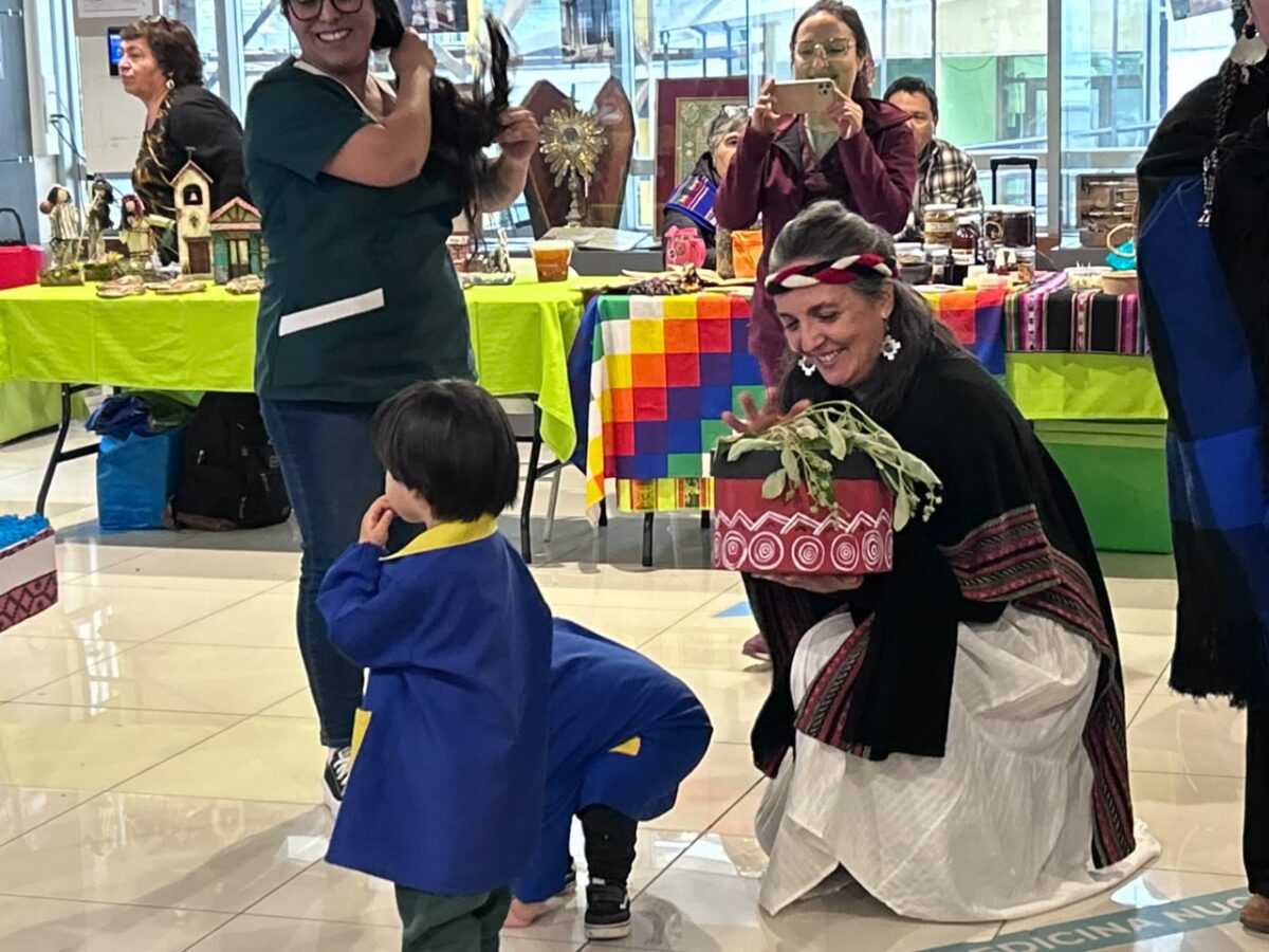 Mujer de pueblo originario agachada mostrando un objeto a un niño en la Feria de Salud Intercultural.