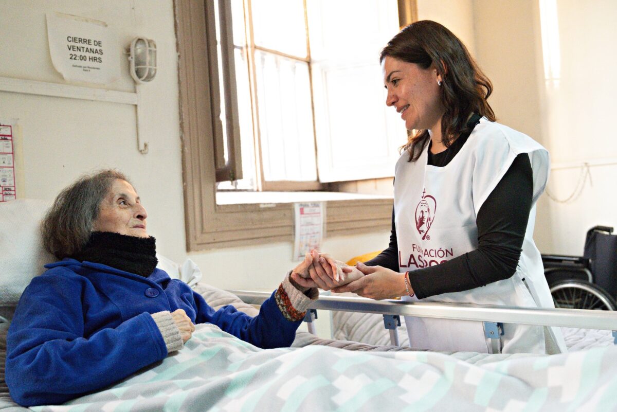 Colaboradora de Fundación Las Rosas tomando de la mano a una adulta mayor en su cama.