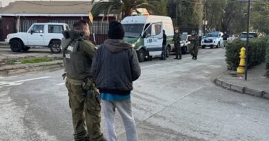 Dos oficiales de Carabineros conversan con un civil en la calle, con vehículos policiales y una furgoneta en el fondo.