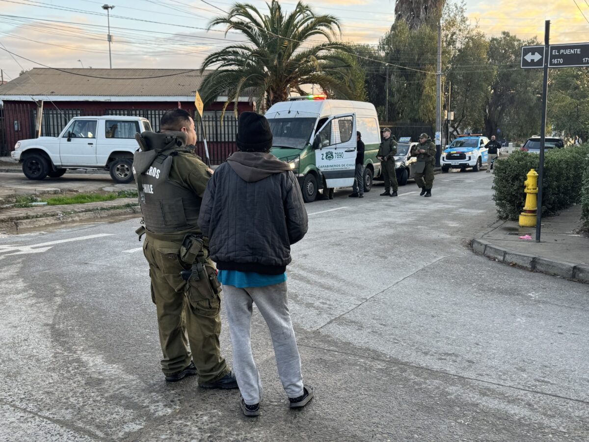Dos oficiales de Carabineros conversan con un civil en la calle, con vehículos policiales y una furgoneta en el fondo.