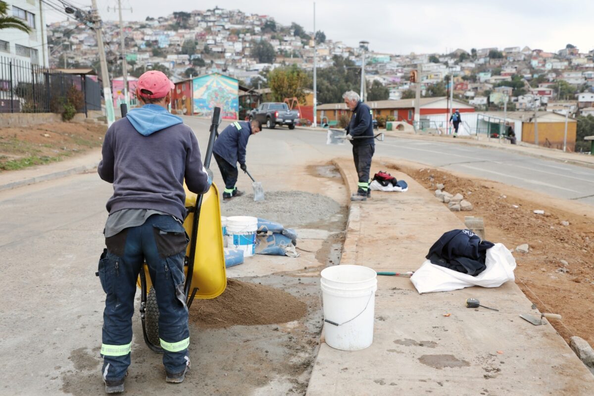 Trabajadores realizan labores de construcción y mejora en un barrio de Valparaíso, con casas asentadas en la ladera de una colina al fondo.