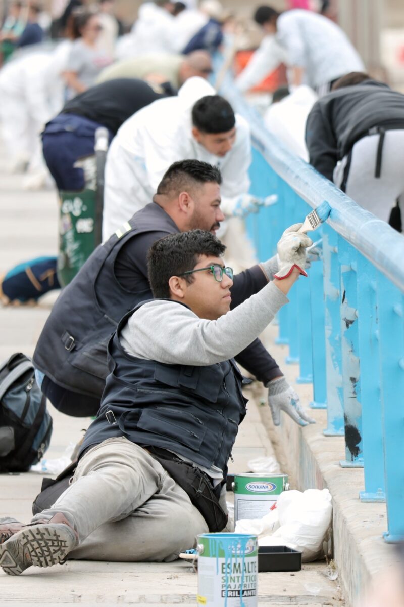 Voluntarios pintan una baranda de color azul brillante como parte de una jornada de mejoramiento urbano.