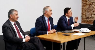 Tres hombres vestidos formalmente sentados en una mesa de madera, con un fondo de pared blanca y ladrillo.