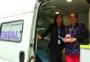 Dos mujeres sonriendo frente a una unidad dental móvil, una sostiene un modelo de dientes.