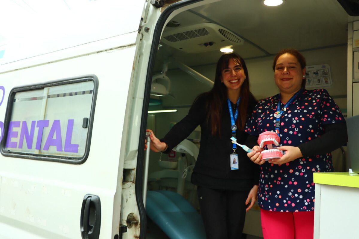 Dos mujeres sonriendo frente a una unidad dental móvil, una sostiene un modelo de dientes.