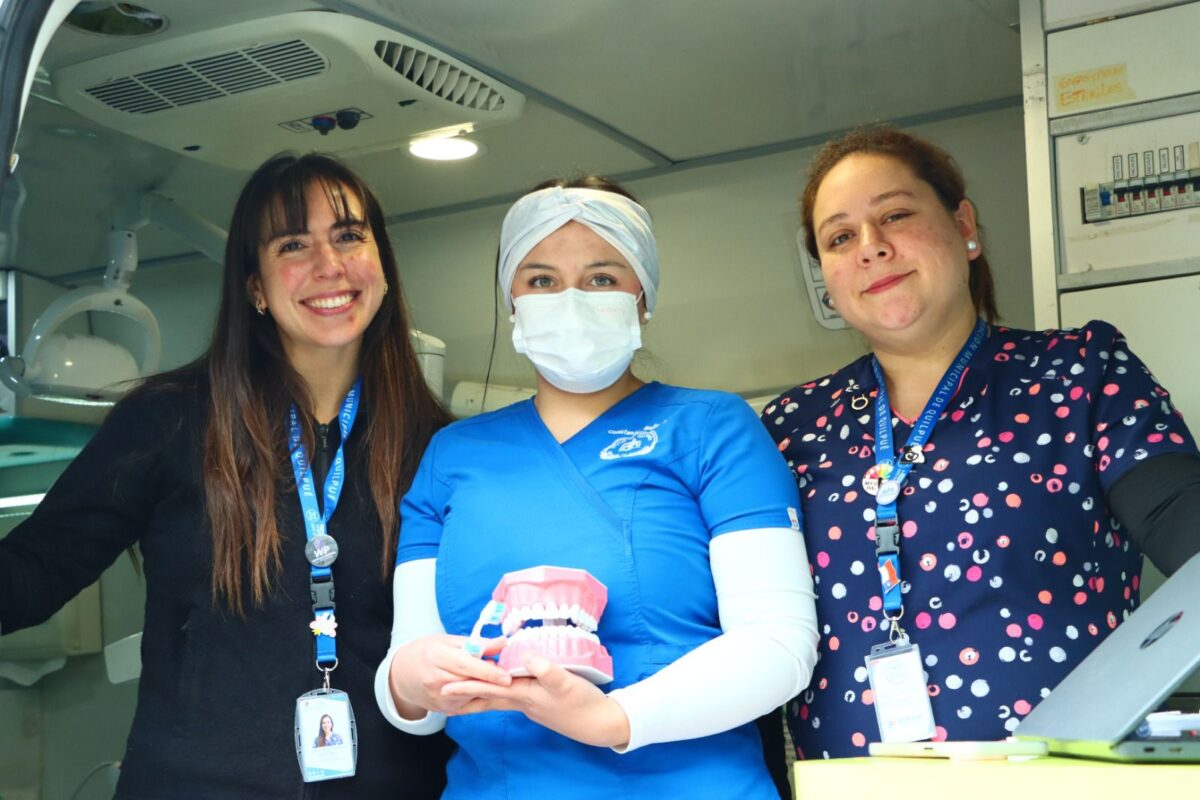 Tres mujeres del equipo de salud dental sonriendo, una sostiene un modelo de dientes.
