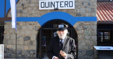 Hombre mayor vestido de uniforme de tren, con un gorro de conductor y un reloj de bolsillo, posando frente a la estación de trenes de Quintero.