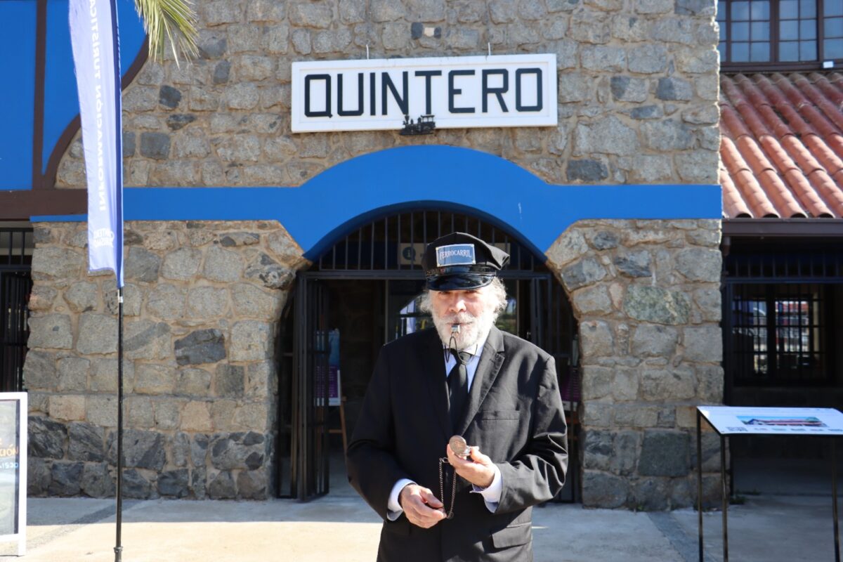 Hombre mayor vestido de uniforme de tren, con un gorro de conductor y un reloj de bolsillo, posando frente a la estación de trenes de Quintero.
