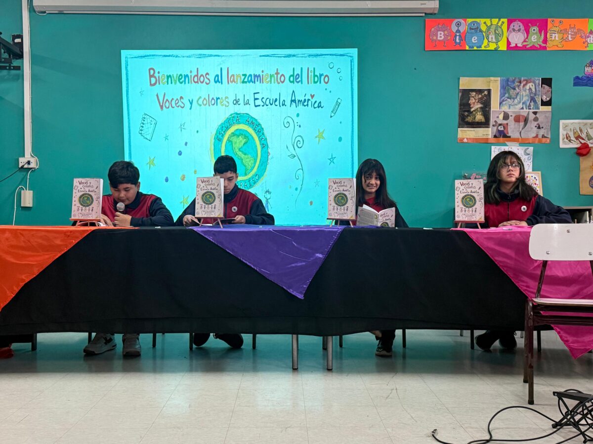 Estudiantes de séptimo básico sentados detrás de una mesa cubierta con manteles de colores, presentando libros.