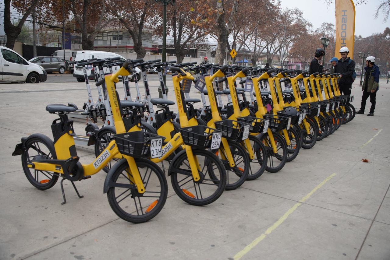 Fila de scooters y bicicletas amarillas de la empresa Whoosh estacionadas en una vereda.