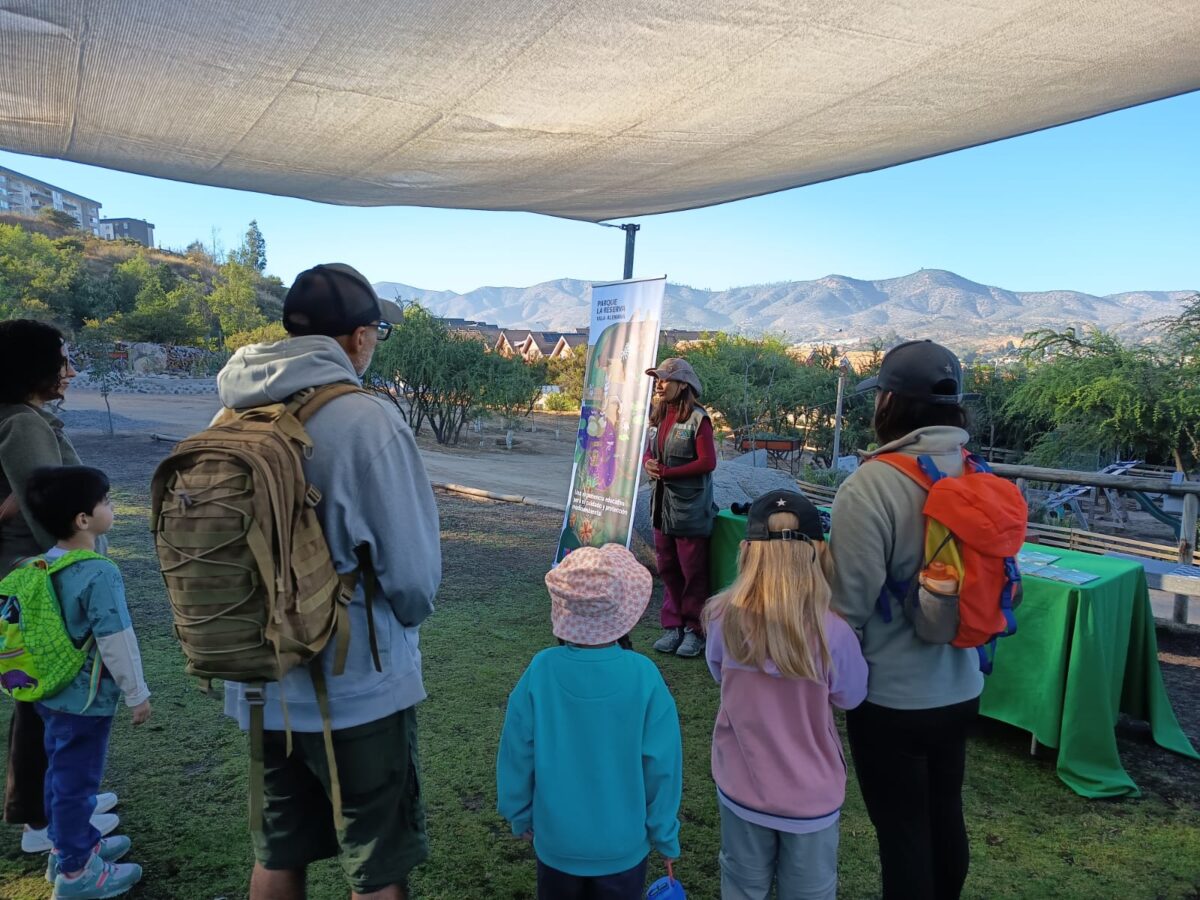 Grupo de personas, incluyendo niños y adultos, observando una presentación al aire libre en un parque con montañas al fondo.