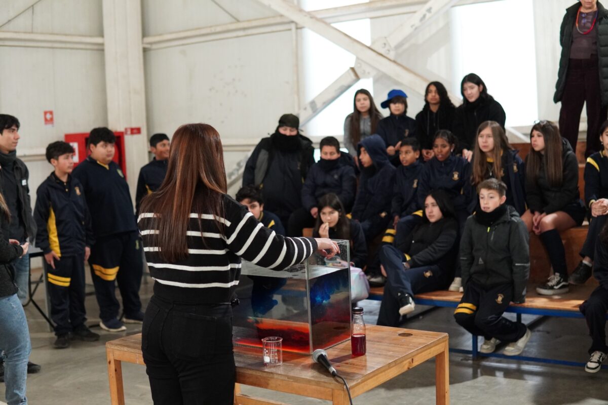 Estudiantes observando un experimento sobre dinámica de fluidos dirigido por una profesora en una sala de clases.